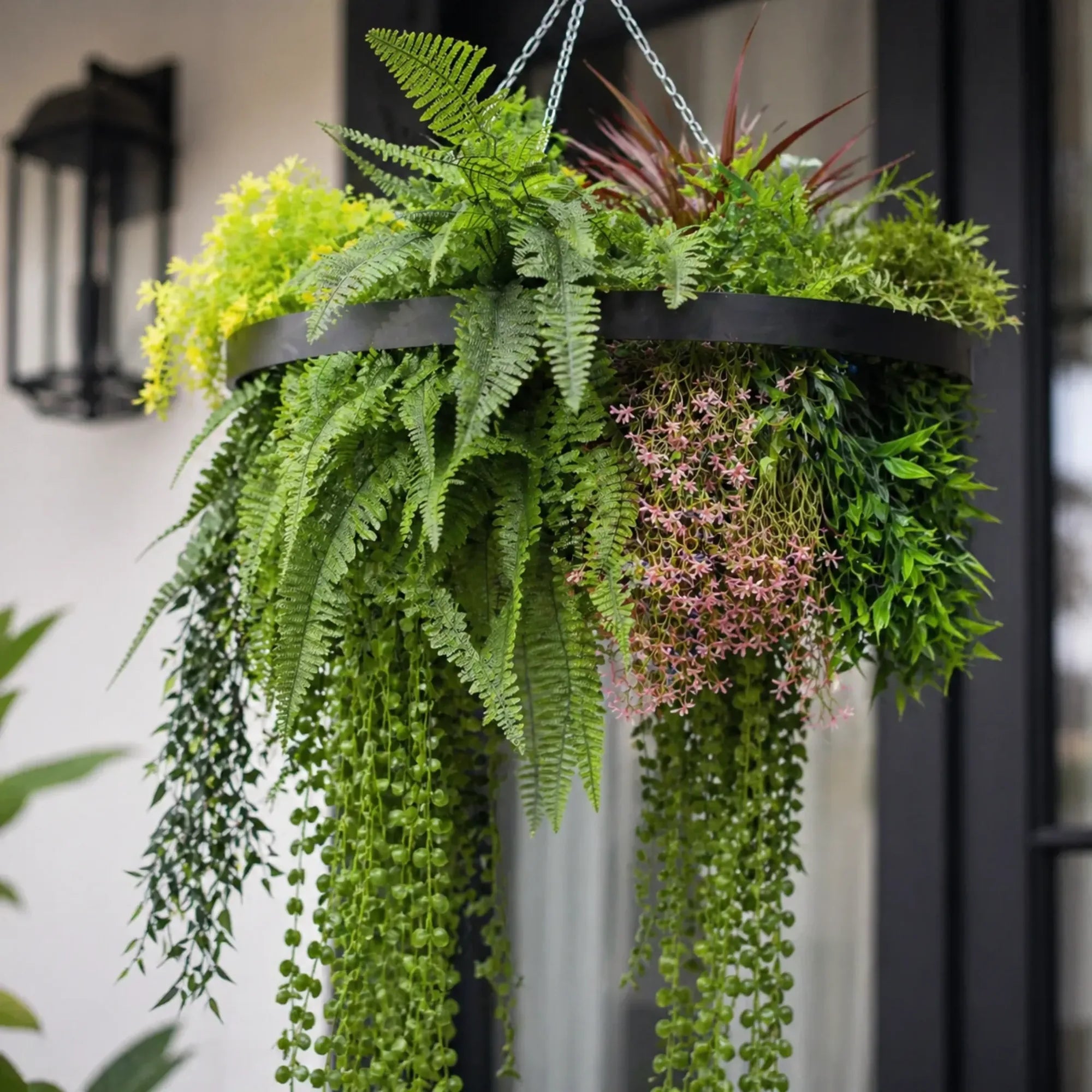 Close-up of a black framed artificial hanging plant chandelier featuring lush tropical foliage, realistic fern leaves, cascading greenery, and delicate pink floral accents.