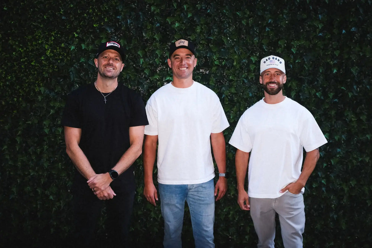 Three men in casual t-shirts and hats standing in front of a green leafy wall.
