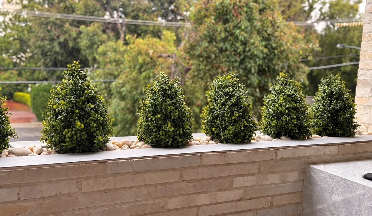 Artificial boxwood topiary plants in a row on a stone windowsill with pebbles.