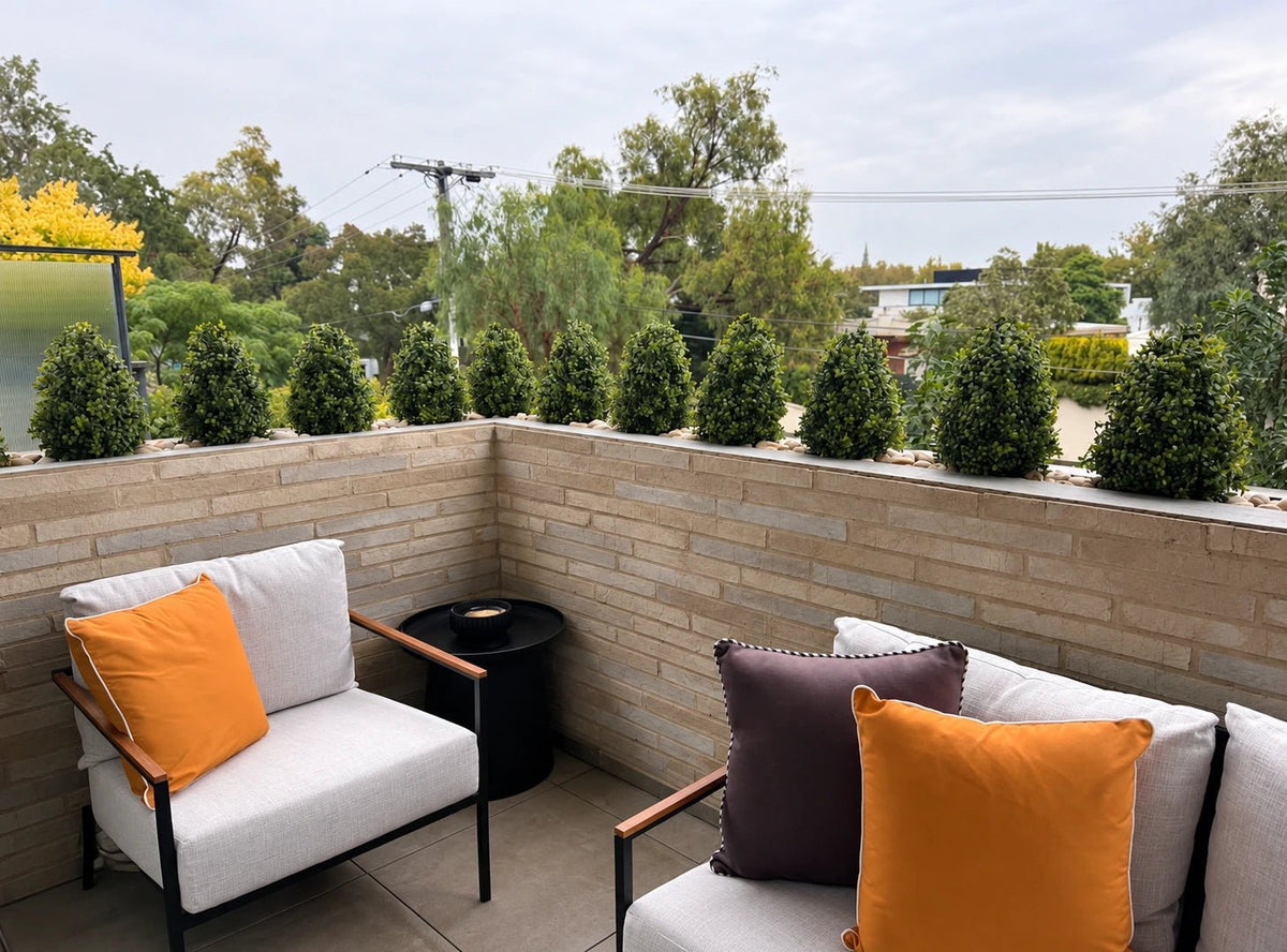 Outdoor balcony with artificial boxwood shrub privacy hedge on brick railing, modern patio chairs.