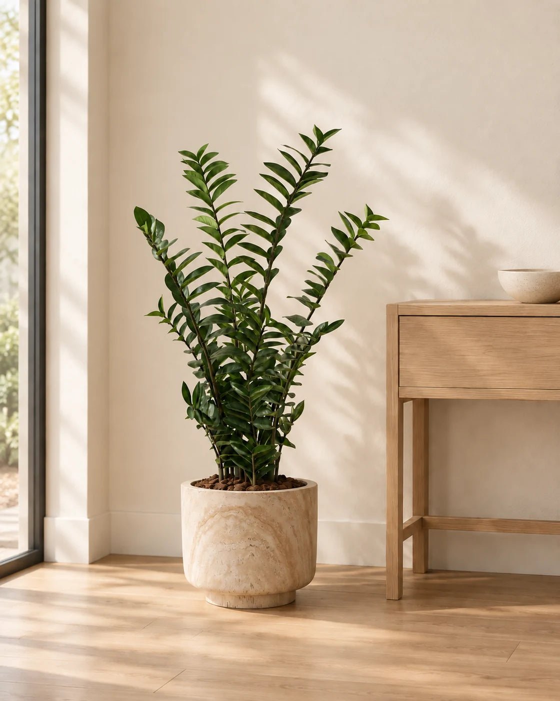 130 cm Zanzibar Gem artificial plant in a marble-effect pedestal planter beside an oak console in a naturally lit interior.
