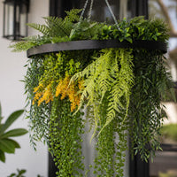 Close-up of an artificial green wall hanging disc with lush mixed foliage, trailing vines, fern leaves, and yellow greenery accents suspended from a circular frame.