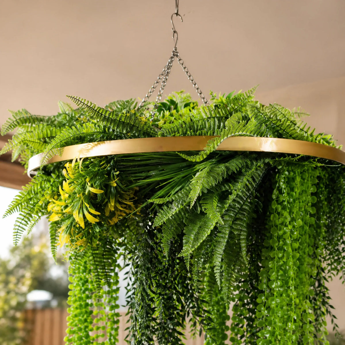 Close-up of a gold framed artificial hanging plant chandelier with draping ferns, cascading greenery, and tropical foliage suspended from a ceiling chain in natural light.