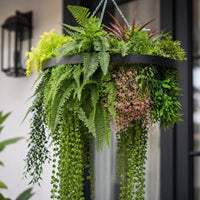 Close-up of a black framed artificial hanging plant chandelier featuring lush tropical foliage, realistic fern leaves, cascading greenery, and delicate pink floral accents.