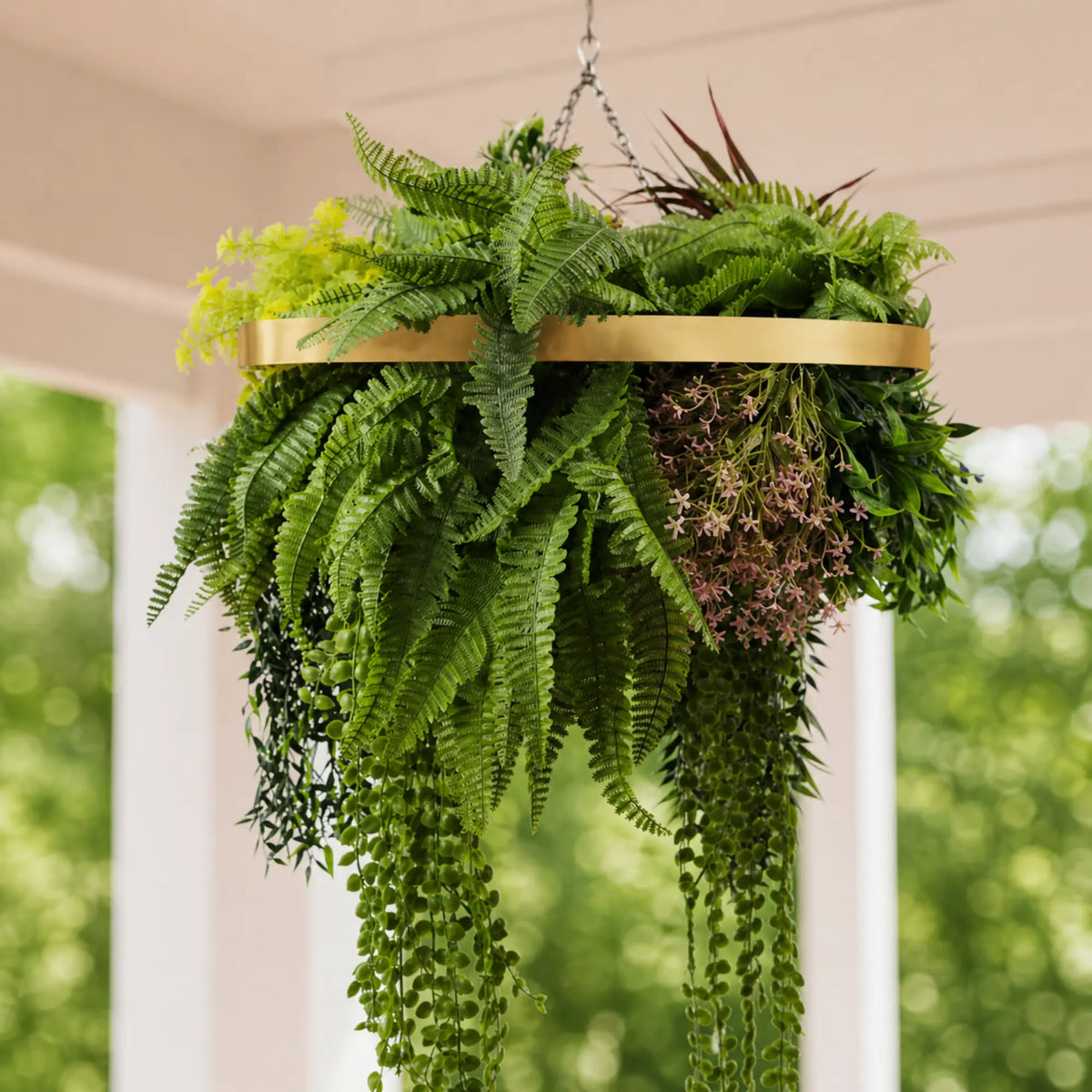Close-up of a gold framed artificial hanging plant chandelier featuring lush tropical foliage, layered fern leaves, trailing greenery, and delicate hanging floral accents suspended by metal chains.