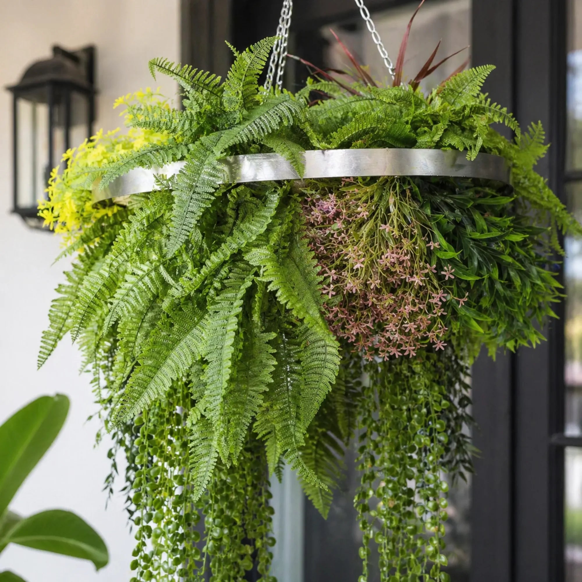 Close-up of a silver framed artificial hanging plant chandelier featuring lush tropical foliage, layered fern leaves, trailing greenery, and delicate floral accents suspended by metal chains.