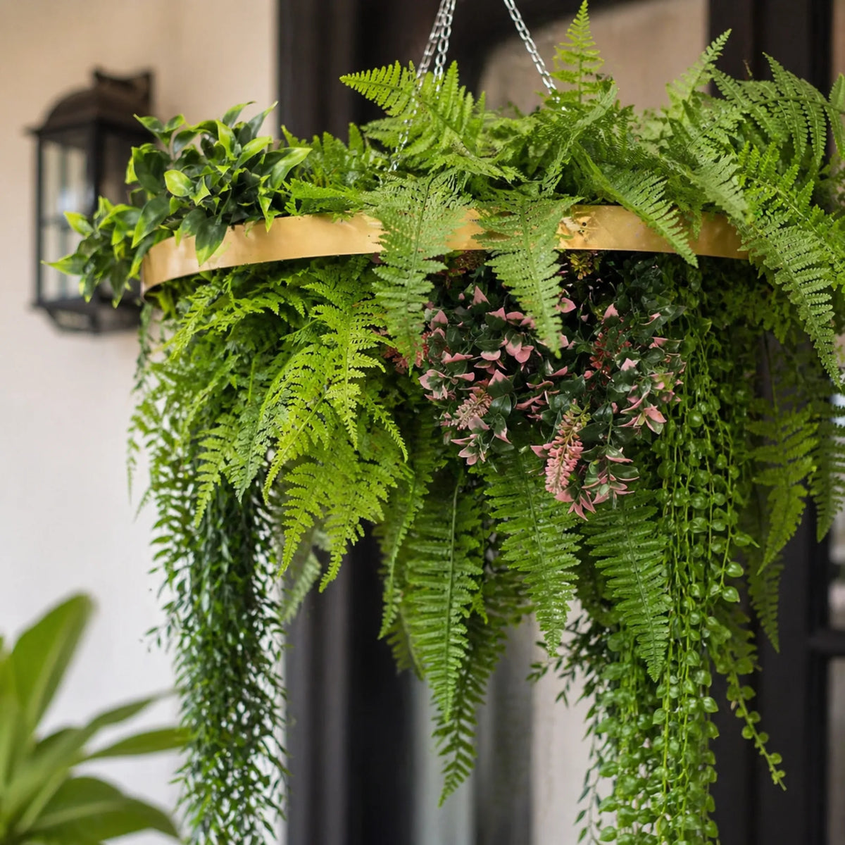 Close-up of a gold framed artificial roof hanging disc with cascading fern foliage, trailing greenery, and pink botanical accents suspended by silver chains.
