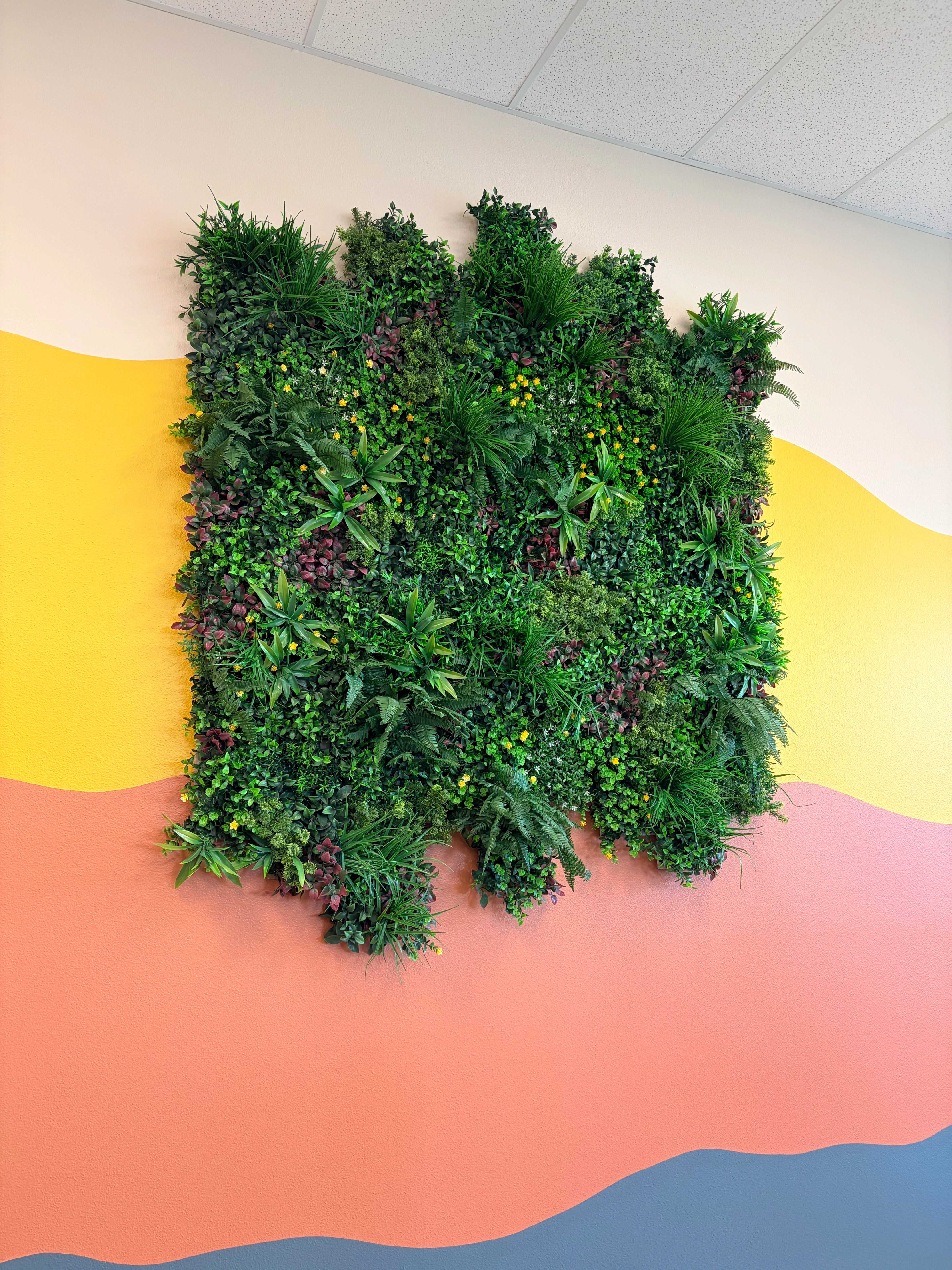 A vertical green wall panel filled with dense artificial plants in different shades of green, including ferns and small flowering accents.