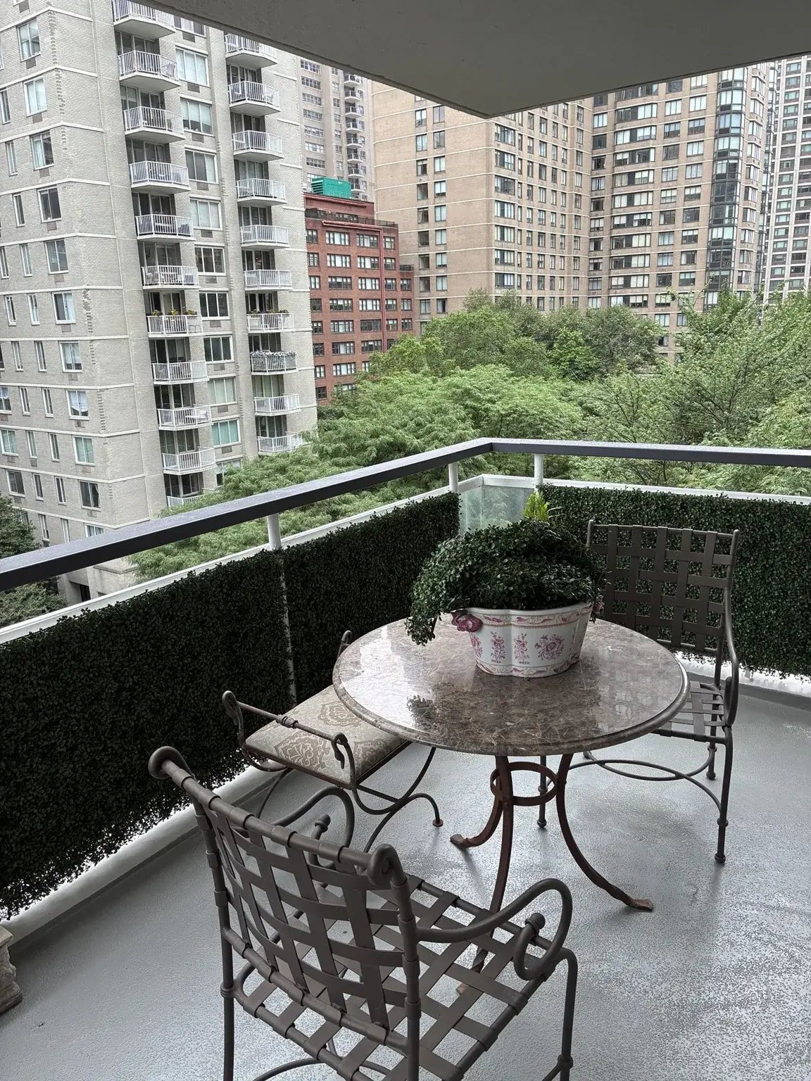 Interior view looking out onto a New York City balcony featuring Dark Boxwood Artificial Vertical Garden Green Wall Panels 40×40in installed along the railing as a privacy screen, with a marble bistro table, iron chairs, and a city skyline backdrop visible through the glass door.