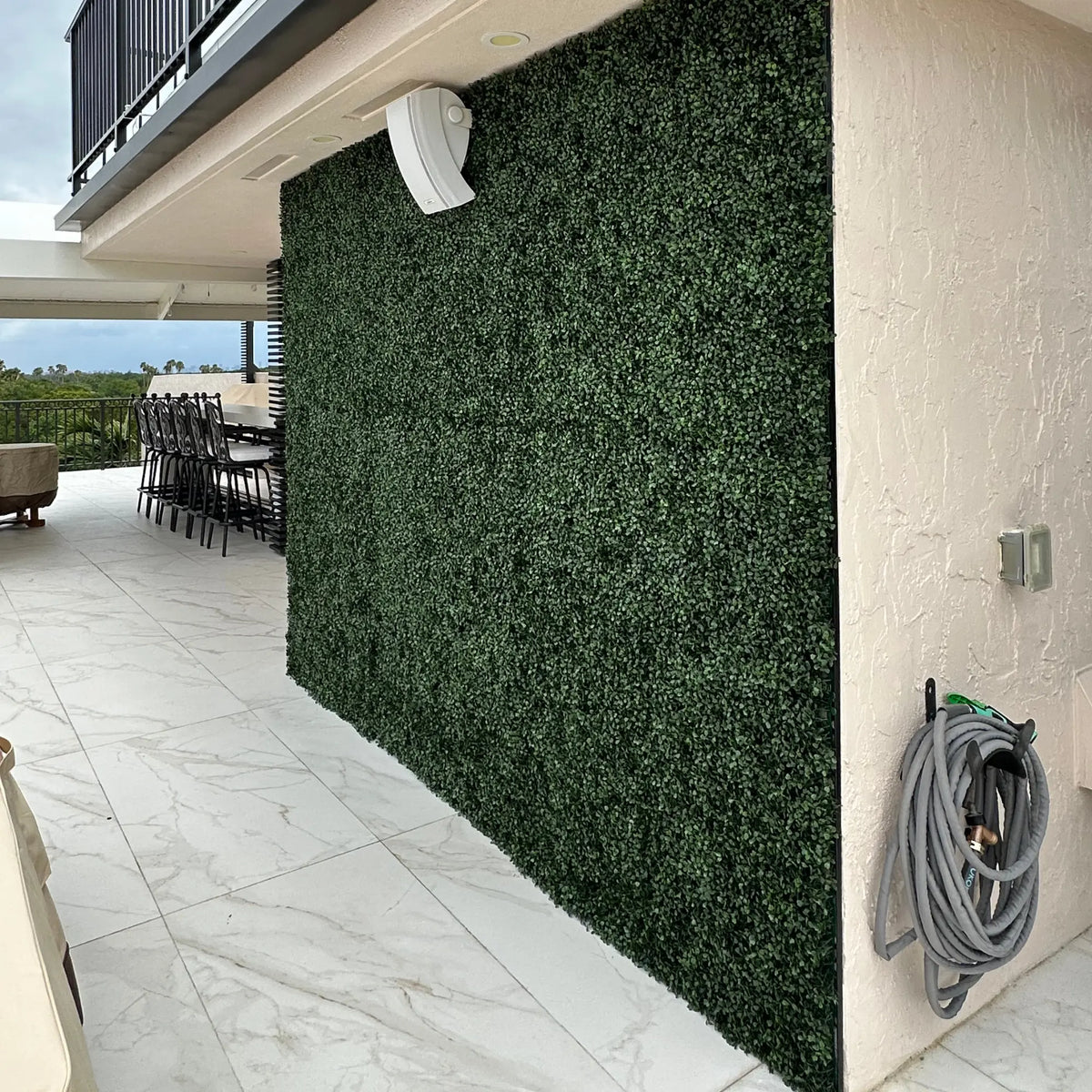 Dark Boxwood Artificial Vertical Garden Green Wall Panel installed along an outdoor entertainment area wall, featuring dense dark green foliage that creates a modern privacy screen beside marble-look tiles and patio seating.