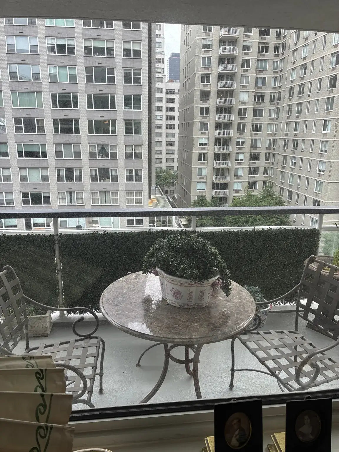 Dark Boxwood Artificial Vertical Garden Green Wall Panel 40×40in 11 SQ FT installed along a balcony railing in New York City, creating a dense, lush green privacy screen with a marble bistro table and iron chairs in the foreground and city buildings surrounded by trees in the background.