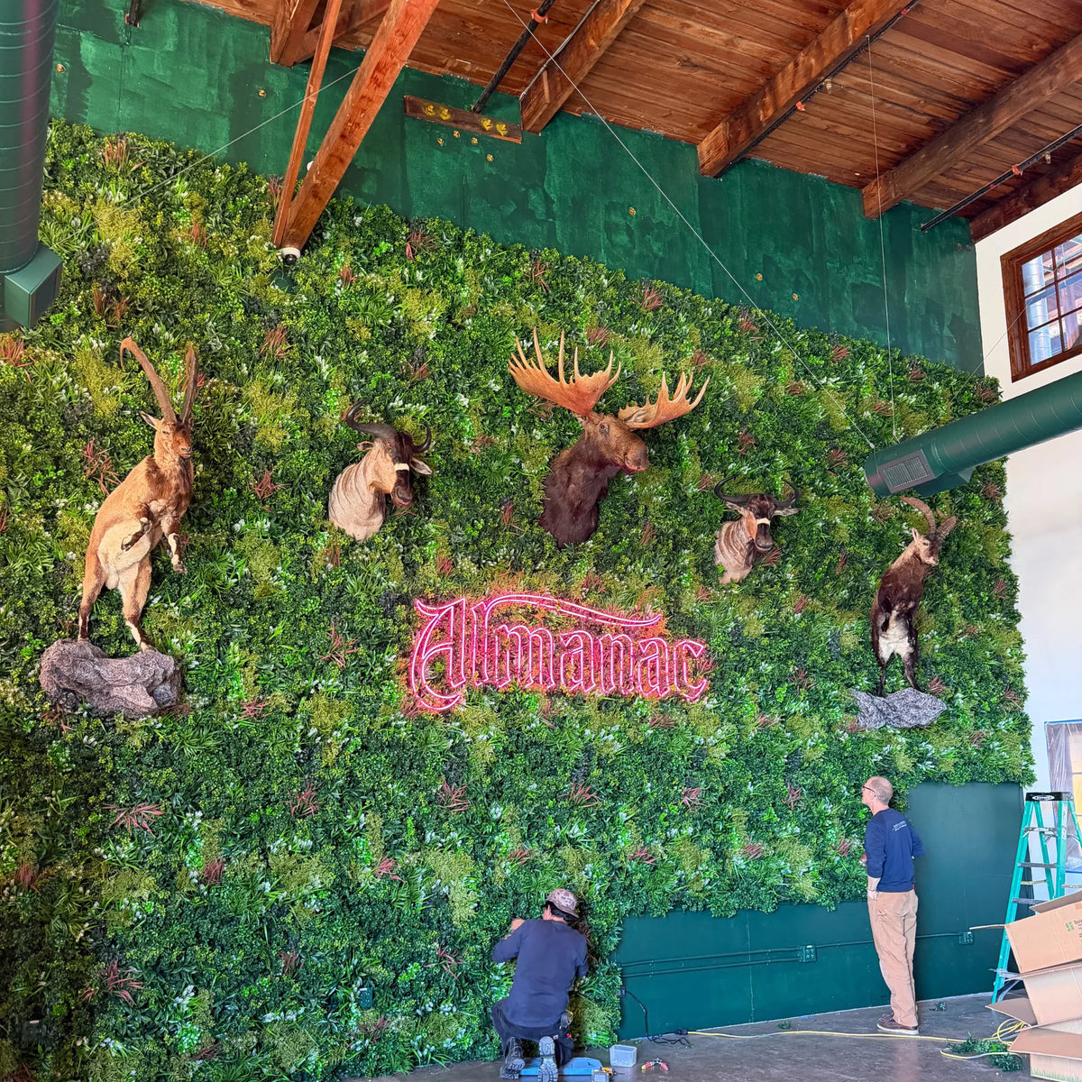 Amazon Jungle Artificial Vertical Garden Green Wall Panel 40×40in 11 SQ FT UV Resistant Commercial Grade installed as a large indoor feature wall with mounted wildlife displays, neon signage, and layered tropical greenery in a commercial venue.