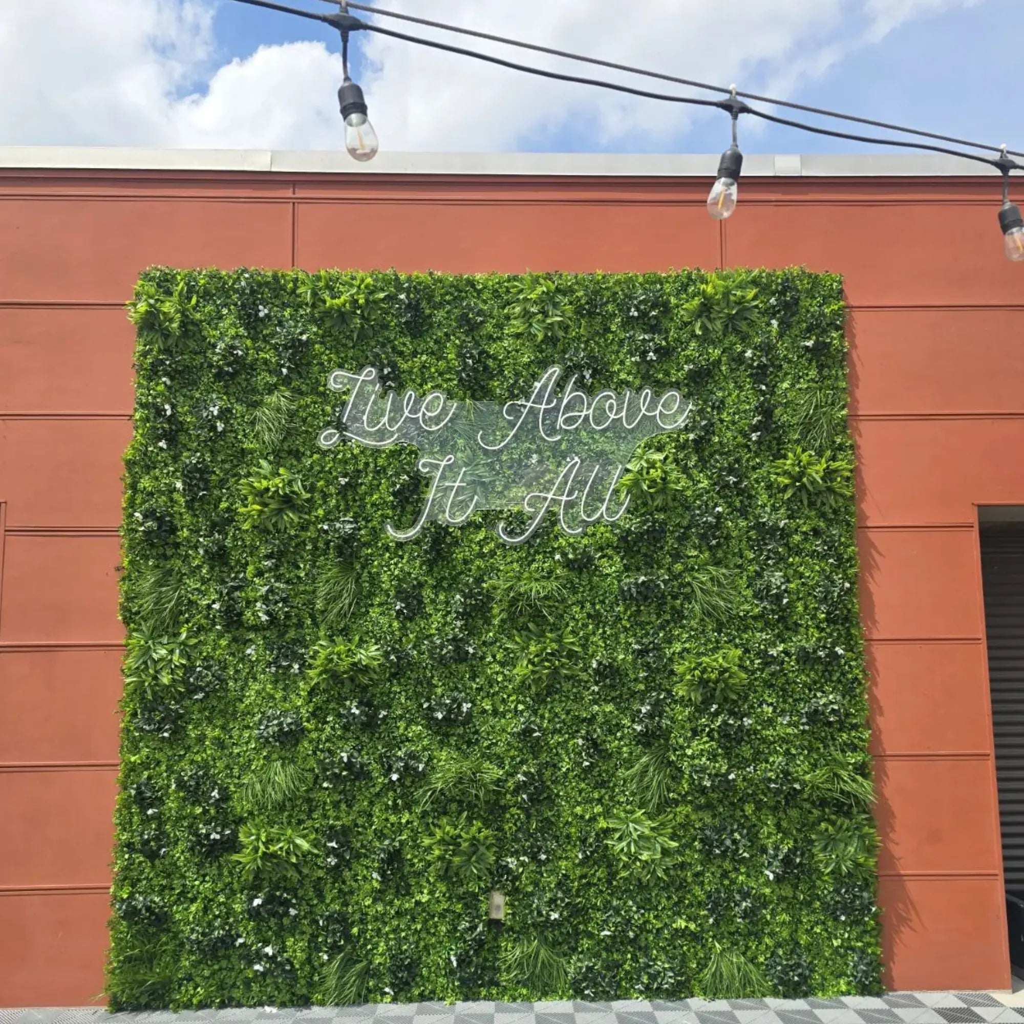 White Oasis artificial vertical garden green wall panels installed as a large commercial outdoor feature wall with layered greenery, decorative lighting, and “Live Above It All” signage on a building exterior.