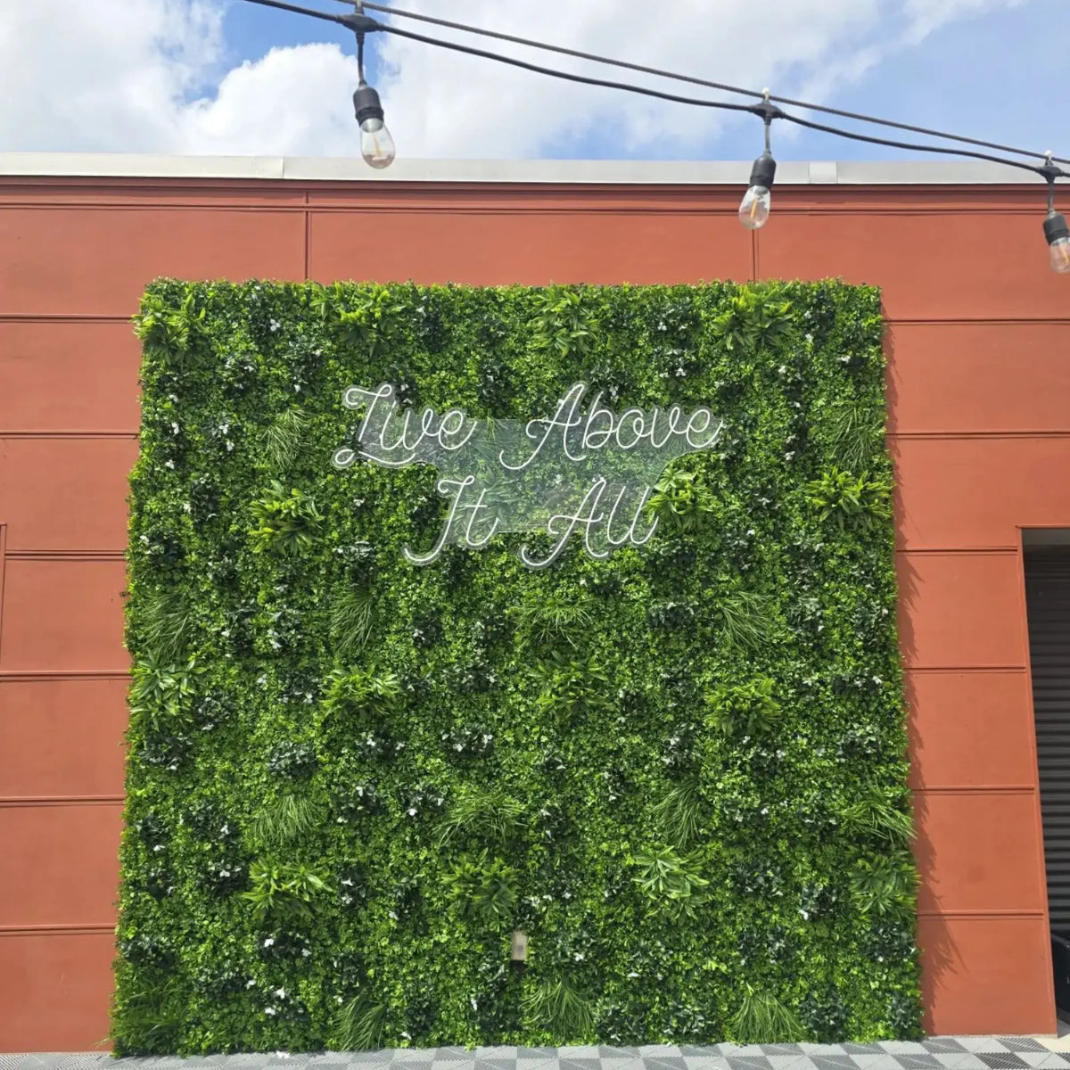 White Oasis artificial vertical garden green wall panels installed as a large commercial outdoor feature wall with layered greenery, decorative lighting, and “Live Above It All” signage on a building exterior.
