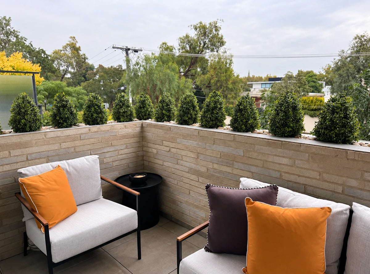 Outdoor balcony with artificial boxwood shrub privacy hedge on brick railing, modern patio chairs.